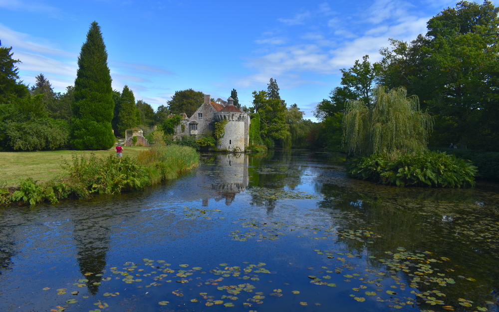 A woman admiring the exterior of a British medieval castle and its reflection in water Exotic gardens with panoramic views of a moated fortress and a Victorian country mansion all in a historic estate