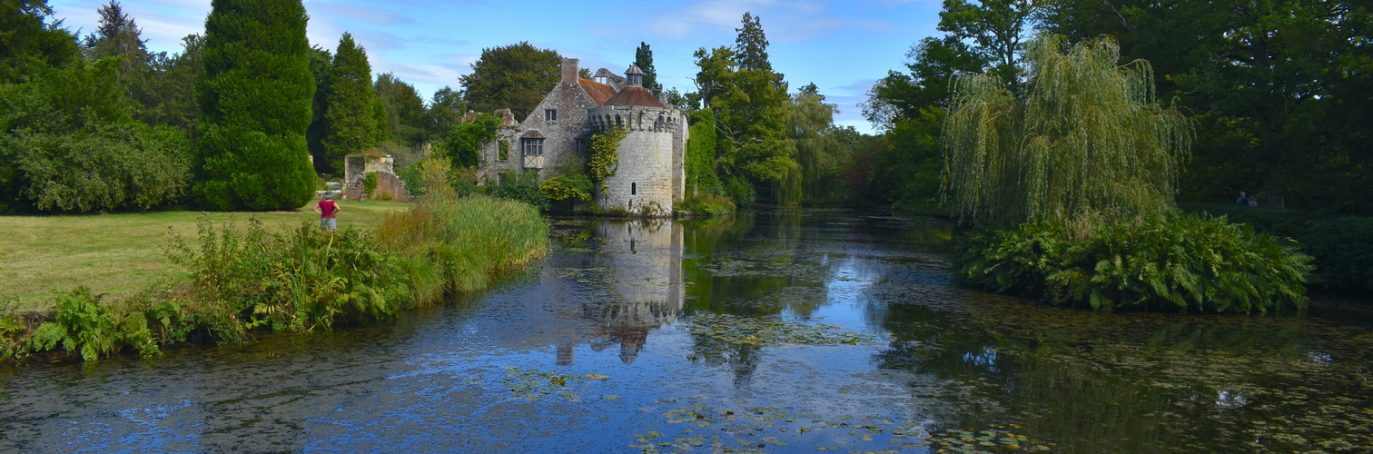 A woman admiring the exterior of a British medieval castle and its reflection in water Exotic gardens with panoramic views of a moated fortress and a Victorian country mansion all in a historic estate