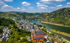 Stadtansicht an einem Fluss, mit Kirche im Vordergrund und Hügeln im Hintergrund.