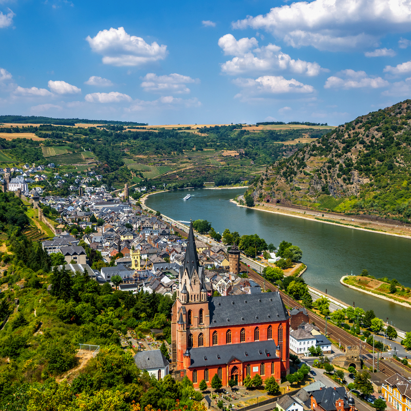 Stadtansicht an einem Fluss, mit Kirche im Vordergrund und Hügeln im Hintergrund.