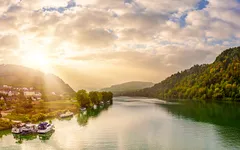 Flusslandschaft mit Bergen, Häusern und Sonnenaufgang im Hintergrund