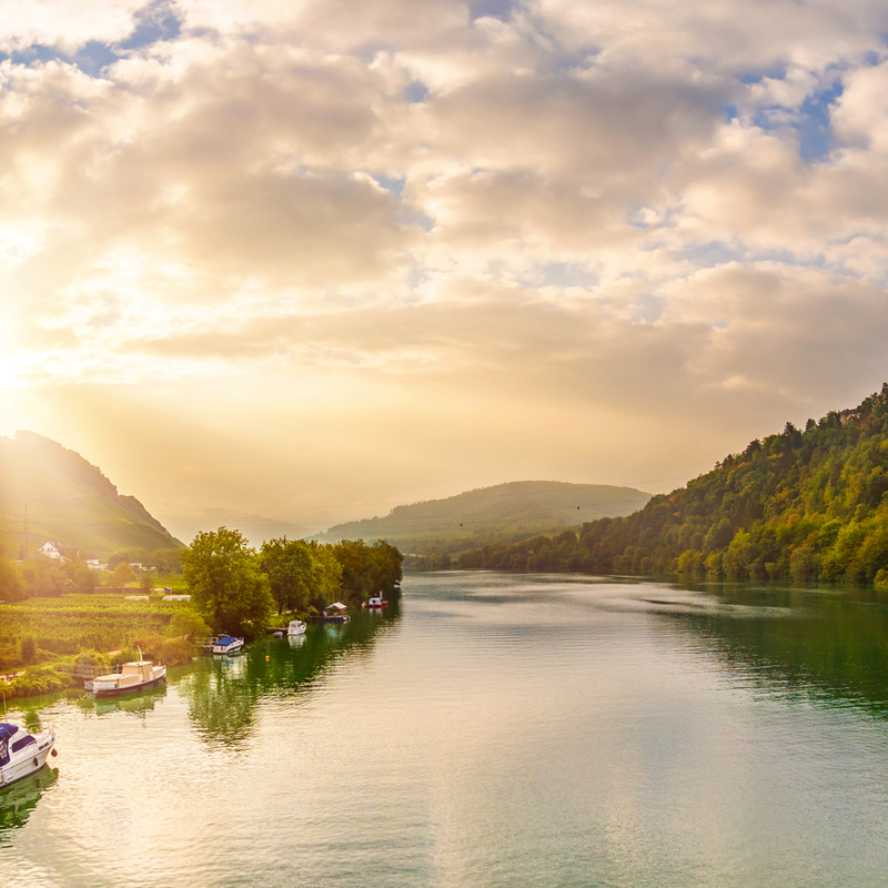 Flusslandschaft mit Bergen, Häusern und Sonnenaufgang im Hintergrund