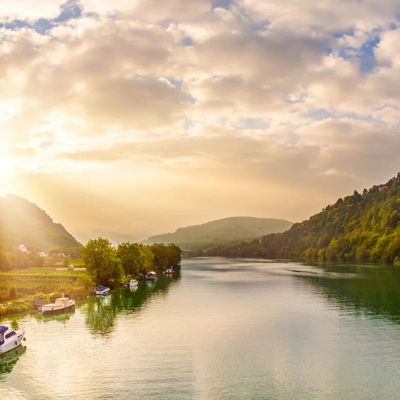 Flusslandschaft mit Bergen, Häusern und Sonnenaufgang im Hintergrund