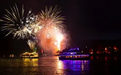 Boote auf einem Fluss bei Nacht mit farbigem Feuerwerk am Himmel.
