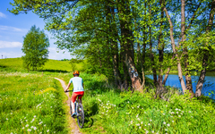 Radfahrerin auf einem Radweg entlang eines Sees und zwischen grünen Wiesen im Landschaftspark Suwalski, Polen