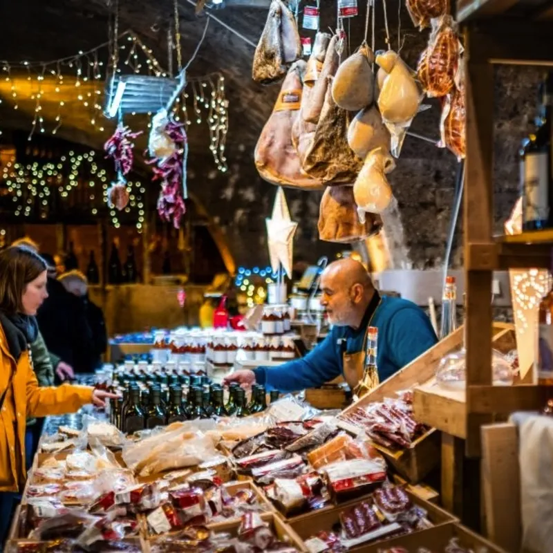 Ein Marktstand mit Wein, Wurst und Käse, besucht von Menschen in einem Keller.