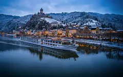 Kreuzfahrtschiff fährt auf einem Fluss vor einer schneebedeckten Stadt mit Hügel im Hintergrund.