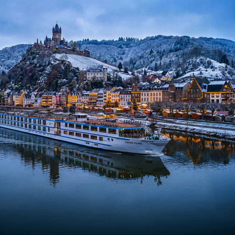 Kreuzfahrtschiff fährt auf einem Fluss vor einer schneebedeckten Stadt mit Hügel im Hintergrund.
