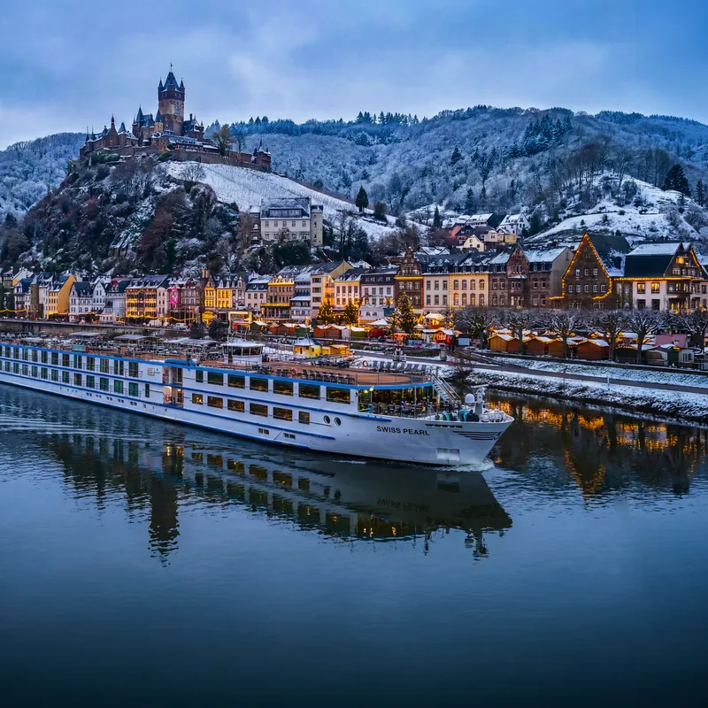 Kreuzfahrtschiff fährt auf einem Fluss vor einer schneebedeckten Stadt mit Hügel im Hintergrund.