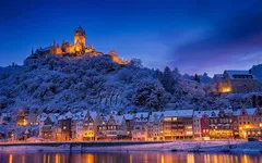 Verschneite Stadt Cochem an der Mosel mit beleuchtetem Schloss bei Abenddämmerung.