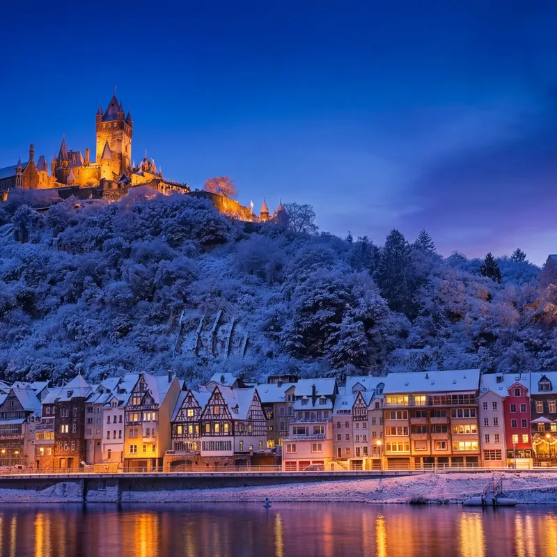 Verschneite Stadt Cochem an der Mosel mit beleuchtetem Schloss bei Abenddämmerung.